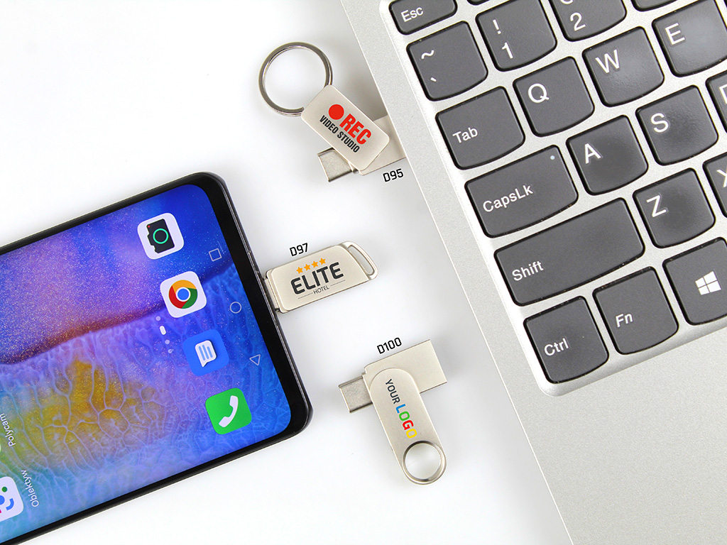 Metalowe pendrive'y 2w1 ze złączami USB-C i USB-A z nadrukowanym logo firmy, zaprezentowane podczas podłączenia do smartfona i laptopa.  